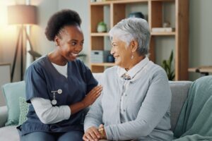 A smiling caregiver in blue scrubs sits beside an elderly woman on a couch, gently touching her shoulder.