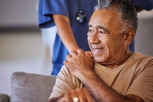 A smiling elderly man sitting comfortably while a healthcare professional places a supportive hand on his shoulder, conveying compassion and care.