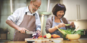 An elderly couple wearing aprons joyfully preparing a healthy meal together in the kitchen, surrounded by fresh vegetables and ingredients.