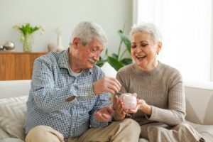 Happy senior couple put coins in piggy bank sit on sofa in living room.