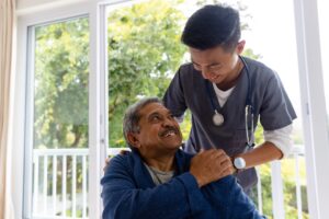 Seated Male Patient in Blue Robe Smiling and Looking Up at Male Healthcare Professional and Holding His Hand at Home